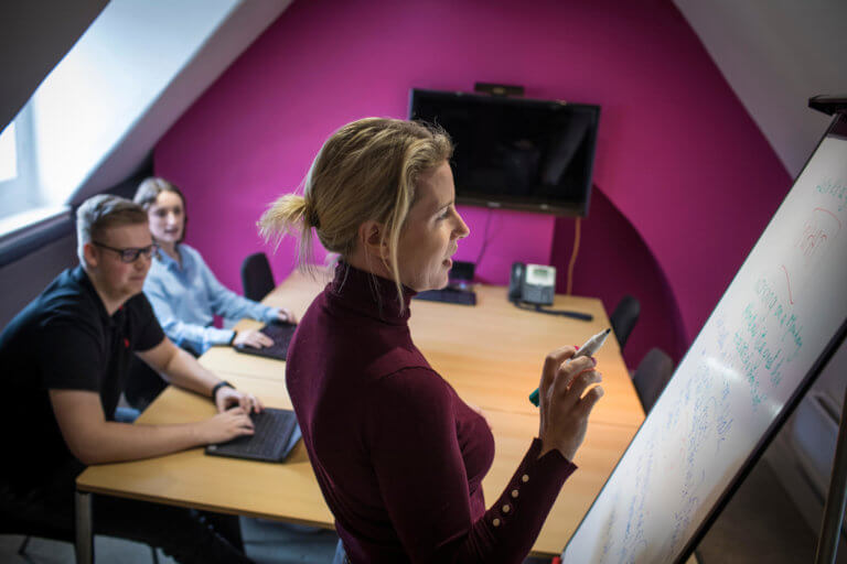 A woman writes on a flipboard in a small conference room while colleagues with laptops watch on